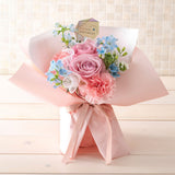 Pastel birthday floral composition with roses, carnation and blue seasonal blooms; greeting label and exact seasonal flowers may differ.
