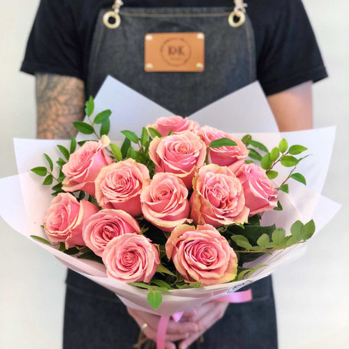 Handheld 12 pink roses with greenery in pale wrap for scale; packaging is illustrative.