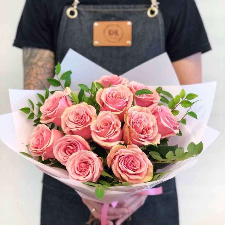 Handheld 12 pink roses with greenery in pale wrap for scale; packaging is illustrative.