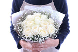 Handheld view of approximately 12 ivory-white roses and baby's breath; reference scale, retail gift wrapping may differ.
